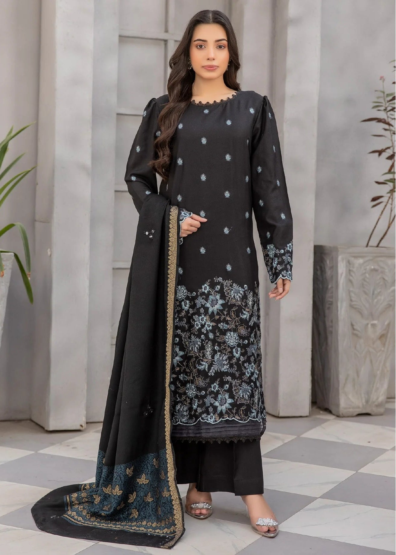 Woman in a black traditional outfit with floral patterns standing in a room with gray walls and a checkered floor.