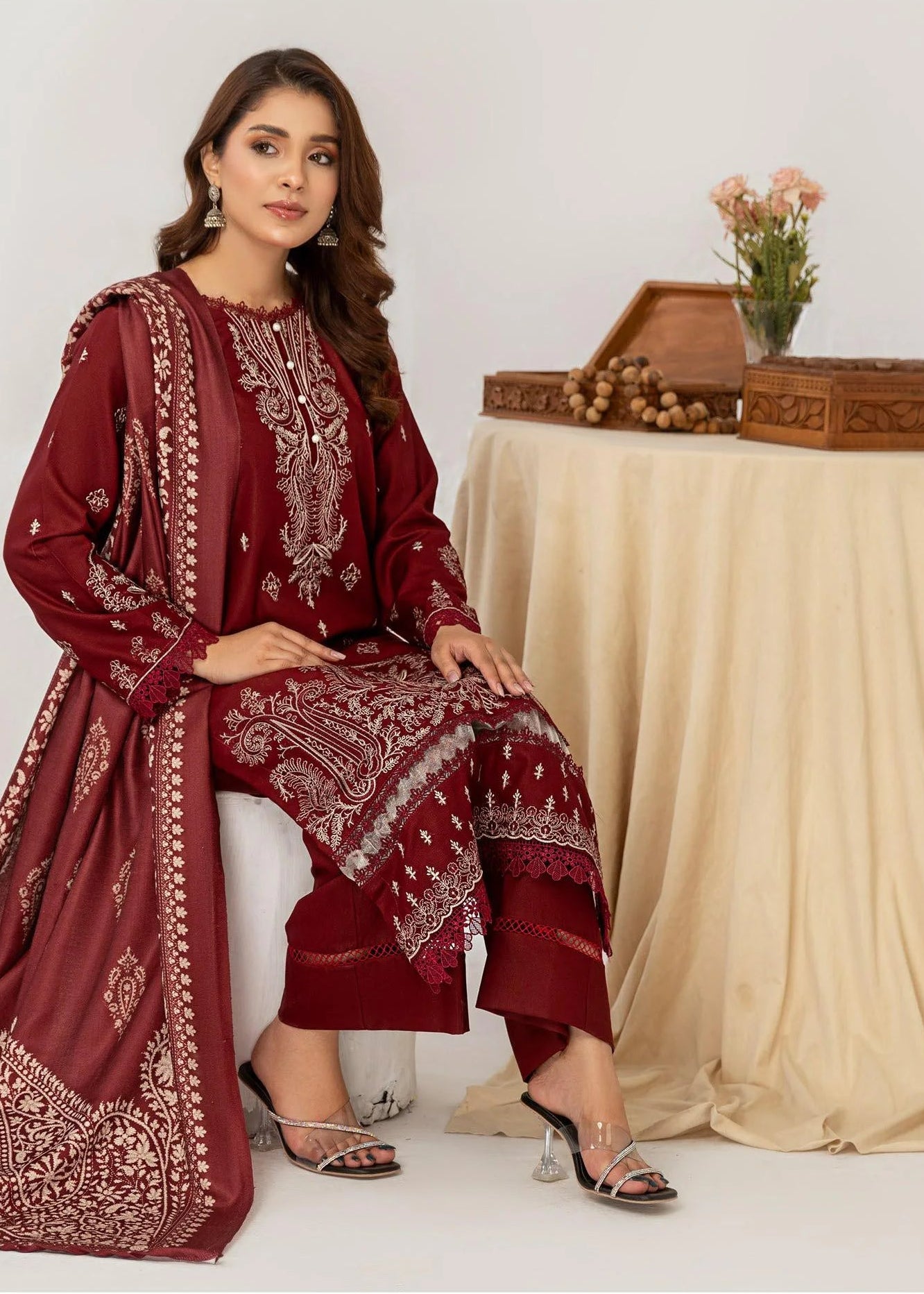 Woman in a red traditional outfit sitting next to a table with a beige cloth and a small plant on a white background
