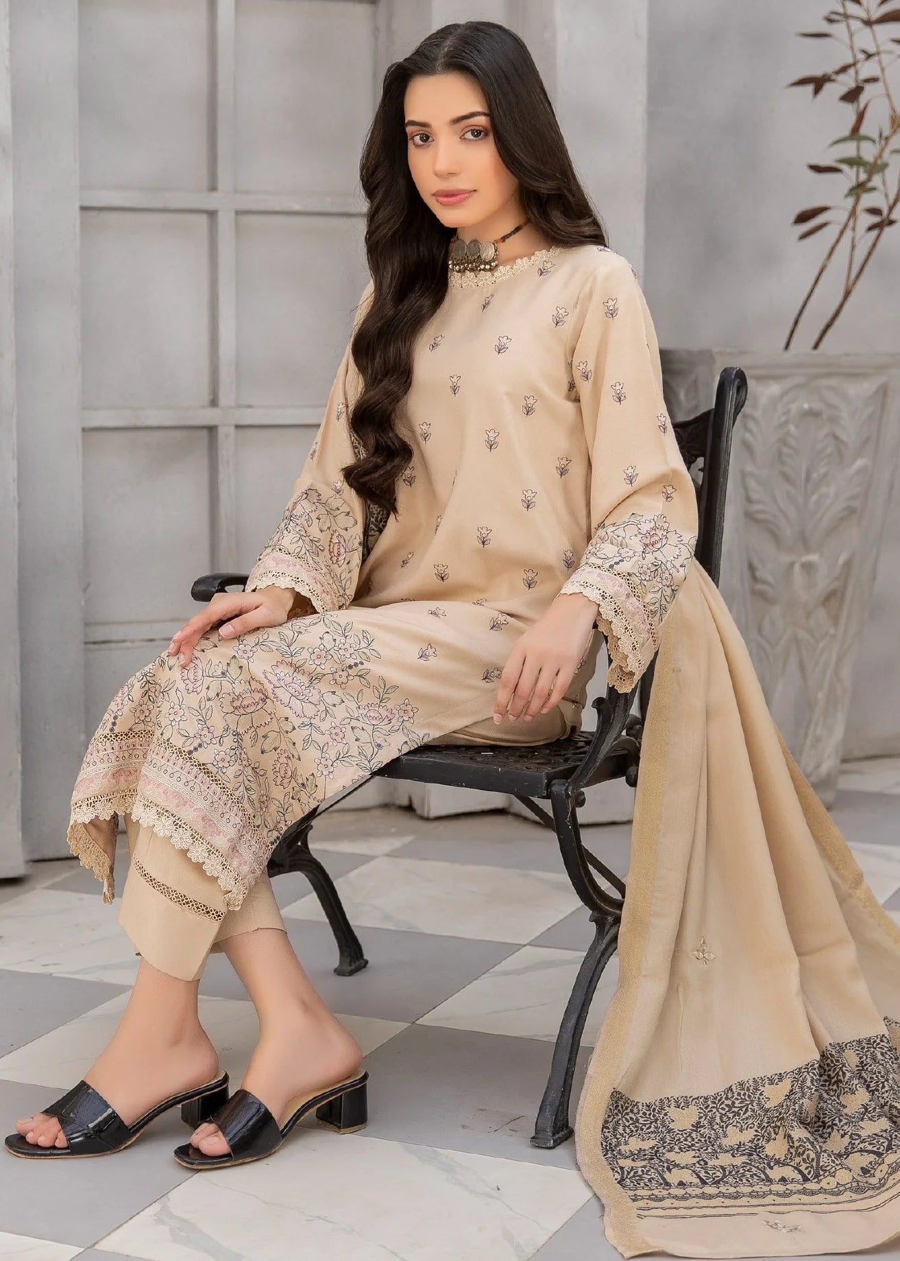 Woman in a beige traditional outfit sitting on a chair in a room with plants and a checkered floor.