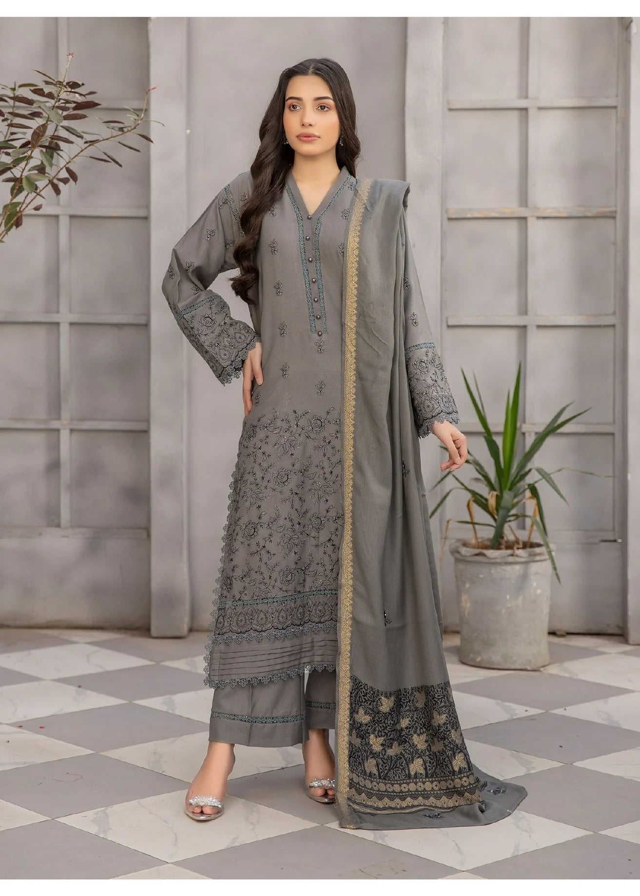 Woman in a gray traditional outfit with floral patterns, standing in a room with decorative elements.