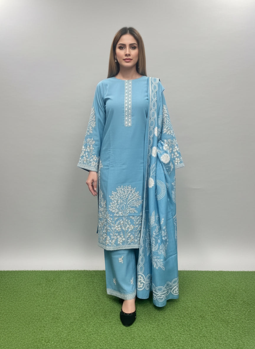 Woman in a blue traditional outfit with white patterns standing against a green textured wall.